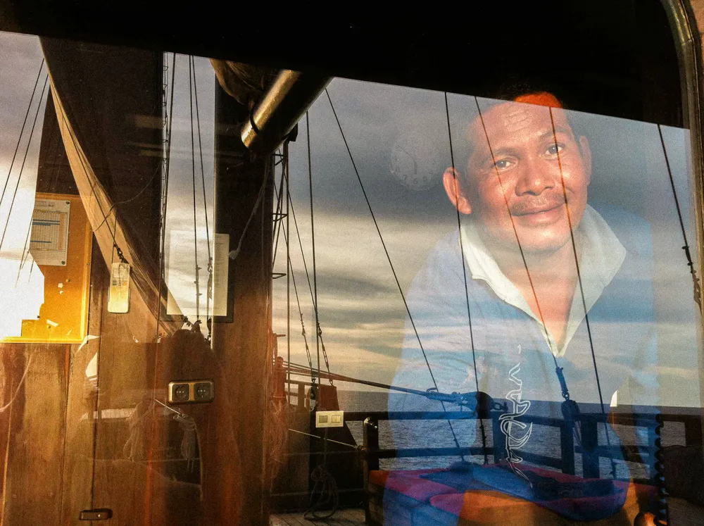 A crew member's face reflected in a window, with the rigging behind.
